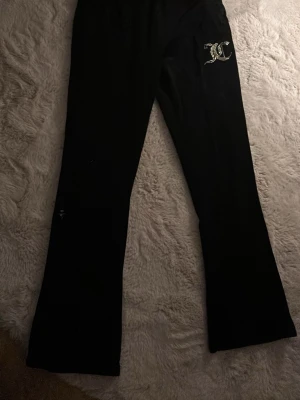 Svarta Juicy Couture mjukisbyxor - Säljer ett par svarta mjukisbyxor från Juicy Couture med glittrande JC-logga i strass på ena höften. Byxorna har utsvängda ben och är gjorda i mjuk velour som känns riktigt skön mot huden. Perfekta för en chill och trendig look. De är knappt använda och har bara en liten fläck som är vit på sidan.