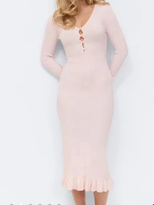 Ljusrosa ribbad långklänning - Supersöt ljusrosa långklänning med ribbad struktur, v-ringning och tre dekorativa knappar framtill. Klänningen har långa ärmar och volangkant nertill för en extra feminin touch. Perfekt för dig som gillar en enkel men ändå elegant stil.