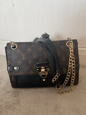 Louis Vuitton monogram axelremsväska - Säljer en klassisk Louis Vuitton handväska i brunt monogrammönster med svarta läderdetaljer och guldfärgad kedja. Väskan har en elegant låsdetalj framtill och justerbar axelrem. Perfekt för dig som vill ha en ikonisk och trendig accessoar.