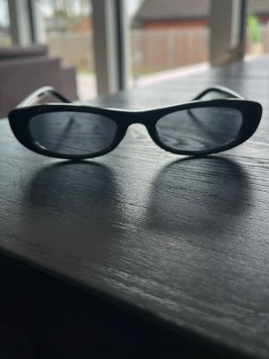 Svarta Saint Laurent solglasögon - Snygga svarta solglasögon från Saint Laurent med klassisk och stilren design. Glasögonen har en rak och något bred båge i blankt syntetmaterial, med Saint Laurent-loggan på sidan. Perfekt för dig som vill ha en ikonisk accessoar.