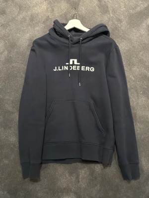 Svart hoodie från J.Lindeberg - Snygg svart hoodie från J.Lindeberg med vit logga på bröstet. Klassisk modell med huva, dragsnören och magficka. Perfekt för en avslappnad och stilren look. Mjuk bomullsblandning som känns skön mot huden.