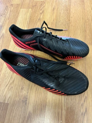 Adidas Predator svarta fotbollsskor - Snygga Adidas Predator fotbollsskor i svart med röda detaljer och vita sulor. Skorna har snörning, mönstrad ovansida och tydliga dobbar för bra grepp på planen. Tillverkade i syntetmaterial och designade för att ge kontroll och precision vid spel.
