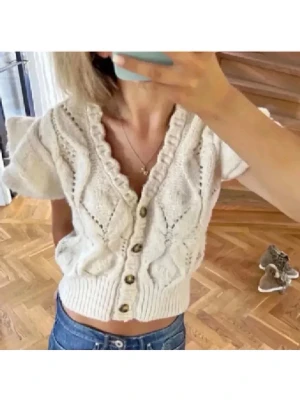 Beige stickad kofta från Zara - Kortärmad beige stickad kofta från Zara med v-ringning, volangkant och dekorativa knappar framtill. Koftan har ett snyggt mönster med struktur och ribbstickad nederkant. Perfekt att styla med jeans eller kjol. Passar flesta storlekar