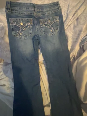 Blå bootcut jeans med dekorerade fickor - Snygga blå bootcut jeans med coola detaljer på bakfickorna, inklusive nitar och mönstrad söm. Jeansen har klassisk femficksdesign och är tillverkade i denim. Perfekta för dig som gillar en avslappnad men trendig stil. Storlek 170