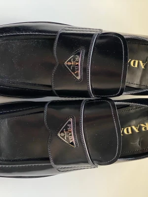 Prada square toe loafer - Snygga svarta loafers från Prada i glansigt lackläder med klassisk fyrkantig tå och dekorativt spänne framtill. Skorna har Pradas ikoniska metallogo på ovansidan och en robust sula som ger en modern touch. Perfekta för dig som gillar exklusiv och stilren design.