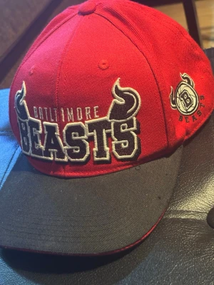 Röd Baltimore Beasts keps från FSBN - Snygg röd keps från FSBN med svart skärm och broderad text 'Baltimore Beasts' framtill. Kepsen har coola horn-detaljer i designen och ett broderat B-märke på sidan. Tillverkad i bomullsmix och har klassisk snapback-form.