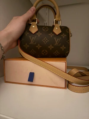 Louis Vuitton Speedy Nano Monogram - Säljer en Louis Vuitton Speedy Nano handväska i klassiskt brunt monogram canvas med beige läderdetaljer och guldfärgade metalldelar. Väskan har både handtag och en lång axelrem, perfekt för dig som gillar lyxiga accessoarer med ikonisk design. Köpte denna på Vinted för 12 tusen med kvitto på bild. Box o dustbag följer med!