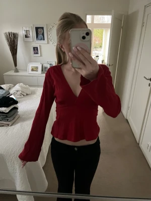Röd omlottopp med volang - Säljer en snygg röd omlottopp med djup v-ringning och volang nedtill. Toppen har långa, vida ärmar och knytdetalj i ryggen. Perfekt för dig som vill ha en trendig och feminin look. Materialet känns mjukt och lätt.