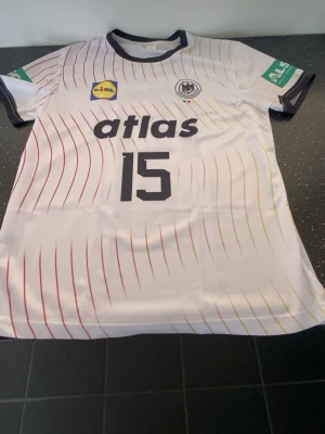 Vit matchtröja med nummer 15 - Vit sporttröja med svarta detaljer och tryck, nummer 15 på framsidan. Tröjan har röda och gula linjer längs hela framsidan, svarta muddar och reklamtryck på bröst och ärm. Perfekt för träning eller match.