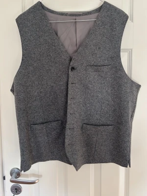 Grå kostymväst från Riley - Stilren grå kostymväst från Riley med klassisk passform. Västen har tre fickor framtill, knäppning med sex knappar och är helfodrad med grått innerfoder. Perfekt att matcha med kostymbyxor för en dressad look. Storlek 54