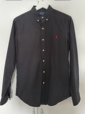 Svart skjorta från Polo Ralph Lauren - Klassisk svart skjorta från Polo Ralph Lauren i slim fit-modell. Skjortan har långa ärmar, knappar framtill och en röd broderad logga på bröstet. Tillverkad i bomull och har button-down krage för en clean look.