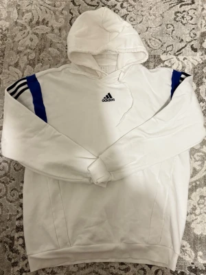 Vit hoodie från Adidas med blå detaljer - Snygg vit hoodie från Adidas med blå ränder på axlarna och svart broderad logga på bröstet. Klassisk huva med snörning och långa ärmar. Perfekt för en sportig och avslappnad stil.