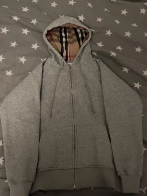 Grå zip-hoodie från Burberry - Snygg grå hoodie från Burberry med dragkedja och klassiskt rutigt foder i huvan. Mjuk bomullskänsla, dragsko i huvan och ribbade muddar. Perfekt för dig som gillar stilrena och exklusiva plagg med en twist.