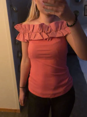 Rosa offshoulder topp med volang - Trendig rosa offshoulder topp med bred volang runt axlarna. Toppen är ribbad och har en figurnära passform som framhäver siluetten. Perfekt för dig som vill sticka ut med en feminin och modern stil.