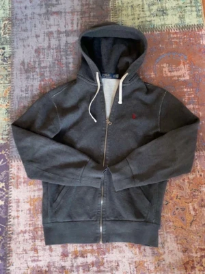 Mörkgrå hoodie från Polo Ralph Lauren - Snygg mörkgrå hoodie med dragkedja från Polo Ralph Lauren. Har vit dragsko i huvan, två fickor framtill och en liten röd logga broderad på bröstet. Perfekt för en chill och stilren look. Tillverkad i mjukt bomullsmaterial.              Storlek S. Hör av dig om du har funderingar! Byten kan vara intressant🙂