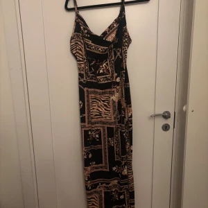 Svart långklänning med mönster slits - Snygg svart långklänning med tunna axelband och unikt mönster i beige och brunt. Klänningen har en figurnära passform och ett coolt mixat print med både rutor och djurmönster. Perfekt för dig som vill sticka ut med en trendig look. Med hög slits