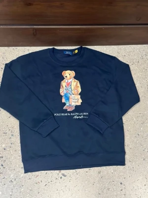 Mörkblå Polo Bear sweatshirt - Mörkblå sweatshirt från Ralph Lauren med ikoniska Polo Bear-trycket framtill. Björnen har på sig jeans, kavaj och håller en shoppingpåse. Tröjan har rund hals och ribbade muddar. Perfekt för en chill och trendig look.
