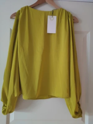 Gul blus med puffärmar - Trendig gul blus med vida, långa puffärmar och resår vid ärmslut. Blusen har en lös passform och är tillverkad i ett lätt, flowy tyg som ger en snygg siluett. Perfekt för dig som vill sticka ut med färg och stil.