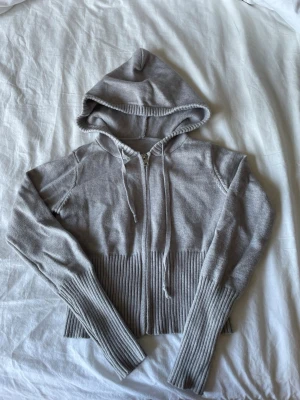 Grå ribbad tröja med dragkedja - Säljer en grå stickad tröja med dragkedja framtill och ribbade detaljer vid midja och ärmslut. Tröjan har huva med dragsko och långa ärmar. Perfekt för en avslappnad och trendig look.