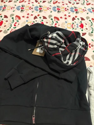 Svart hoodie från Burberry med dragkedja - Svart hoodie från Burberry med klassiskt rutigt foder i huvan och dragkedja framtill. Broderad Burberry-logga på bröstet och ribbade muddar vid ärmar och midja. Fick i present men säljer vidare då den är för stor. passformen är något stort i storlek men väldigt snygg. Nypris: ca 8000