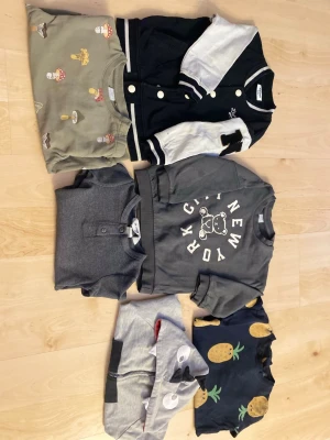 Mix av barntröjor och hoodie - Säljer ett paket med sex olika barntröjor och en hoodie. Innehåller t-shirts med svamp- och ananastryck, en grå sweatshirt med björn och text, en svartvit baseballjacka, en grå piké och en grå hoodie med djurmotiv. Flera olika färger och roliga tryck.