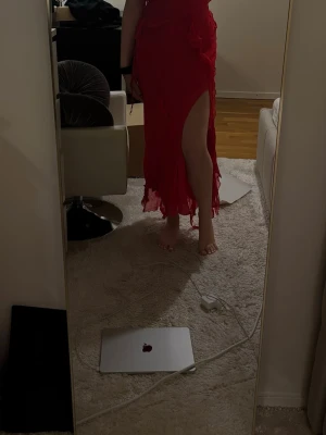 Röd långklänning med slits - Säljer en röd långklänning med hög slits och volangdetaljer. Klänningen har omlottmodell och markerad midja med knytband. Perfekt för dig som vill sticka ut med en elegant och feminin look.