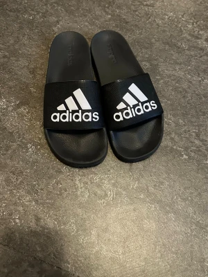 Svarta Adidas badtofflor - Säljer ett par klassiska svarta Adidas badtofflor med vitt logotryck på remmen. Tofflorna har en enkel slip-on design och är tillverkade i syntetmaterial som tål vatten. Perfekta för stranden, gymmet eller hemma. Det står storlek 47 men passar bättre 45/46