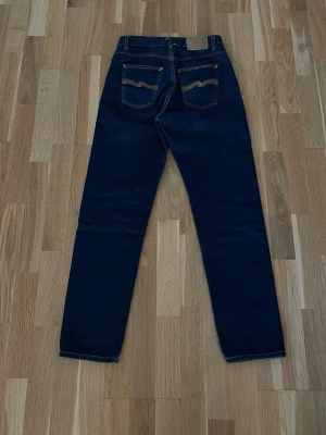 Mörkblå jeans från Nudie Jeans - Snygga mörkblå jeans från Nudie Jeans, Modell:Breezy Britt, med raka ben och klassisk femficksmodell. Jeansen har kontrastsömmar och ikoniska bakfickor med vågigt broderi. Tillverkade i kraftig denim, perfekta för en tidlös look.                                   Pris är inte fast🙂