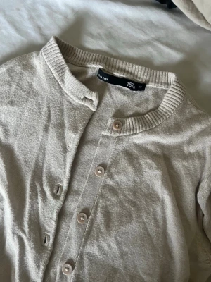 Beige ribbad kofta med knappar - Ljusbeige kofta med rund halsringning och ribbade muddar. Koftan har långa ärmar och stängs med matchande knappar framtill. Perfekt att slänga över en t-shirt eller linne för en enkel och clean look.