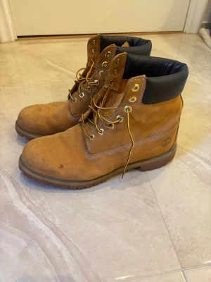 Klassiska Timberland  - Säljer ett par ikoniska Timberland kängor i ljusbrun nubuck med svart vadderad kant och grov sula. Kängorna har guldfärgade öljetter och gula snören. Perfekta för dig som vill ha en tidlös och street-inspirerad look. 