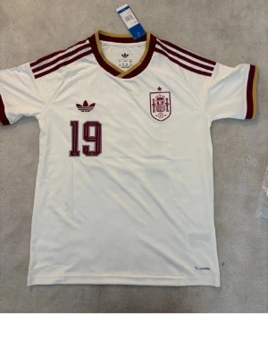 Vit Spanien fotbollströja Adidas - Säljer en vit fotbollströja från Adidas med Spaniens landslagsemblem och nummer 19 i vinrött. Tröjan har vinröda detaljer på axlar och ärmslut samt gul krage. Tillverkad i lätt och ventilerande material, perfekt för träning eller match.