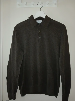 Mörkbrun stickad polotröja - Mörkbrun stickad tröja med krage och tre knappar framtill. Tröjan har lång ärm och ribbade muddar vid ärmslut och nederkant. Perfekt för dig som gillar en stilren och klassisk look.