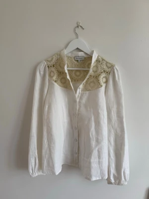 Vit blus med spetsdetaljer från Dea Kudibal - Stilren vit blus med vackra spetsdetaljer i beige över axlar och bröst. Blusen har knäppning framtill, klassisk krage och långa ärmar med resår vid ärmslut. Tillverkad i ett lätt och luftigt tyg.
