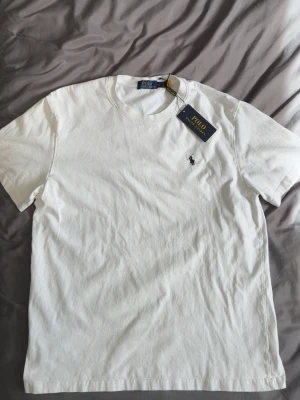 Vit t-shirt från Polo Ralph Lauren - Vit t-shirt från Polo Ralph Lauren med klassisk rund hals och korta ärmar. T-shirten har den ikoniska broderade loggan på bröstet och är gjord i mjuk bomull. Perfekt för en clean och stilren look. XXL FÖR BARN ÄR L FÖR HERR