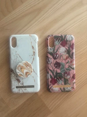 Två mobilskal från iDeal of Sweden - Säljer två snygga mobilskal från iDeal of Sweden. Ett har vit marmordesign med popsocket i matchande mönster, det andra är rosa med blommigt motiv. Båda är hårda skal i plast och passar modeller med kameraöppning uppe till vänster.