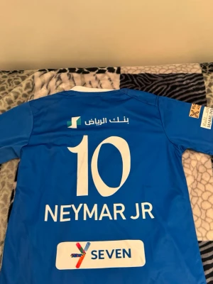 Blå Al Hilal Neymar Jr matchtröja - Snygg blå matchtröja från Al Hilal med Neymar Jr och nummer 10 på ryggen. Tröjan har vita detaljer vid kragen och tryck från flera sponsorer. 