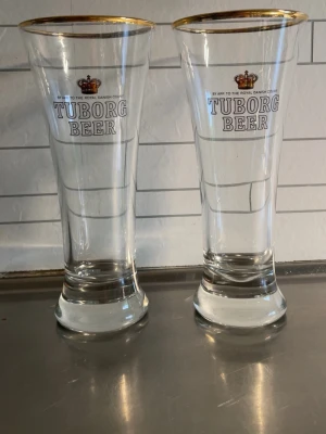 Tuborg Beer ölglas med guldkant 80kr st - Två höga ölglas från Tuborg Beer med guldkant och tryckt logga samt krona. Glasen är genomskinliga och har en klassisk ölglasform med bred fot och smalare topp. Perfekta för att servera öl med stil.