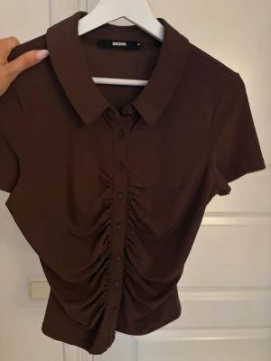 Brun kortärmad topp från Bik Bok - Snygg brun topp från Bik Bok med korta ärmar och krage. Toppen har knappar framtill och en cool rynkad detalj längs knappraden som ger en trendig look. Perfekt till jeans eller kjol för en stilren outfit.