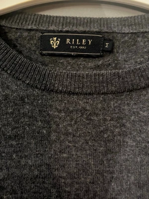 Mörkgrå stickad tröja från Riley - Mörkgrå stickad tröja från Riley med rund halsringning och långa ärmar. Tröjan har en klassisk, enkel design och ribbstickade muddar vid hals, ärmslut och nederkant. Perfekt för lager på lager och passar till det mesta.
