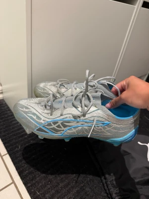 Silvriga Puma fotbollsskor - Säljer ett par riktigt snygga silvriga fotbollsskor från Puma med blå detaljer och klassisk snörning. Skorna har en låg profil och är tillverkade i ett lätt syntetmaterial som ger bra bollkontroll och speed på planen. Perfekta för dig som vill sticka ut på matchen! Använda en endaste gång därav i fint skick. Inga slitage eller defekter. Finns KVITTO