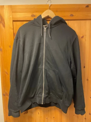 Svart zip hoodie med fickor - En enkel svart hoodie med dragkedja framtill och två stora fickor. Modellen har huva med dragsko och ribbade muddar vid ärmar och nederkant. Perfekt för en avslappnad streetwear-look.