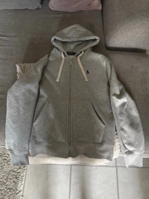 Grå zip hoodie från Polo Ralph Lauren - Snygg grå hoodie med dragkedja från Polo Ralph Lauren. Klassisk design med två fickor fram, vit dragsko i huvan. Mjuk bomullsblandning för extra komfort. Tröjan är i storlek S men är mindre i storleken så om man har storlek XS så passar den perfekt.