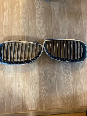 BMW njurgrillar i svart och grå original - Säljer ett par BMW njurgrillar i svart plast med kromad ram. Passar till bilens front och har den klassiska vertikala ribbdesignen. Perfekt för dig som vill byta ut eller uppgradera grillen på din BMW.