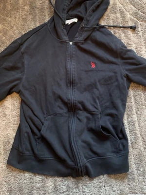 Svart hoodie från U.S. Polo Assn - Svart hoodie med dragkedja från U.S. Polo Assn. Klassisk design med röd broderad logga på bröstet, huva med dragsko och känguruficka framtill. Tillverkad i mjukt bomullsmaterial som känns skönt mot huden. Perfekt för en avslappnad och stilren look. Liten k storleken tröjan är också lite skadad,så fråga efter bilder i förväg