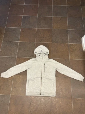 Ljusgrå Sail Racing hoodie med dragkedja - Ljusgrå hoodie med hel dragkedja framtill och två fickor. Tröjan har en huva med snörning och ett diskret tryck på bröstet. Tillverkad i mjukt sweatshirtmaterial, perfekt för en avslappnad stil. Passar både till jeans och mjukisbyxor.