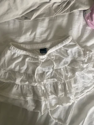 Vit volangkjol med snörning - Supersöt vit kortkjol från Gina Tricot med flera volanger och snörning i midjan. Ser väldigt liten ut men den är stretchig! Knappt använd. 