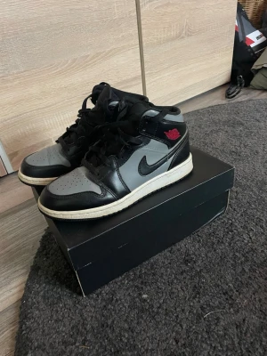 Air Jordan 1 Mid svart/grå - Säljer ett par Air Jordan 1 Mid i svart och grått läder med klassisk Swoosh på sidan och röd Air Jordan-logga vid ankeln. Skorna har svart snörning, perforerad tåbox och vit sula. Perfekta för dig som gillar streetwear och sneakerskultur. Skorna har heeldrag bak. Priset är inte fast!