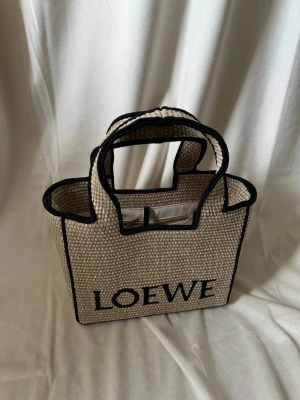 Beige handväska  - Snygg beige handväska  i flätad canvas med svarta kanter och logga framtill. Väskan har dubbla handtag och en öppen design med innerfack. Perfekt för dig som gillar stilrena accessoarer med en lyxig touch.