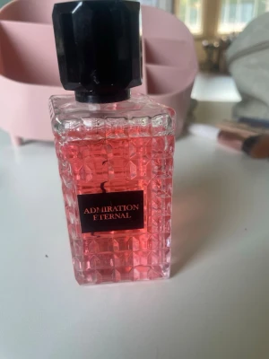 Admiration Eternal parfym rosa glas - Parfymflaska i fyrkantigt, mönstrat glas med rosa innehåll och svart etikett där det står 'Admiration Eternal'. Flaskan har en stor svart, åttakantig kork som ger ett lyxigt intryck. Perfekt för dig som gillar snygga detaljer på sminkbordet.