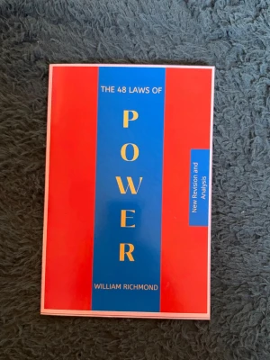 The 48 Laws of Power bok - Säljer boken 'The 48 Laws of Power' av William Richmond. Omslaget är rött och blått med gul text. Boken är i pocketformat och har en modern design. Perfekt för dig som vill lära dig mer om maktspel och strategier.